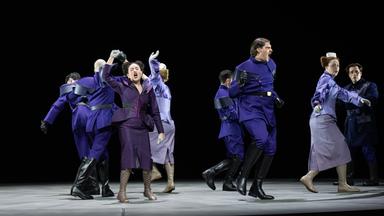 Bühnenszene: Menschen in violett-farbener Bekleidung tanzen auf einer Bühne.