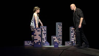 Bühnenszene: Eine Frau und ein Mann stehen sich gegenüber und sehen sich an. Zwischen ihnen steht auf dem Boden das Modell einer Stadt.