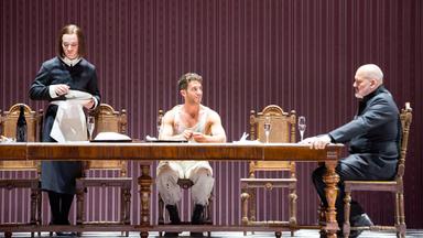 Bünenszene: Ein junger Mann mit weißer, blutbespritzter Bekleidung sitzt an einer Tafel. Am Ende der Tafel rechts, sitzt ein älterer Mann. Eine Hausangestellte, links im Bild, räumt das Geschirr ab.