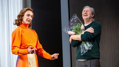 Bühnenszene: Eine Frau in orange-farbenem Kleid steht neben einem Mann, der einen Blumenstrauß in den Armen hält.