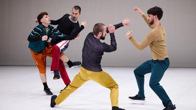 Bühnenszene: Vier Männer in bunter Bekleidung tanzen.