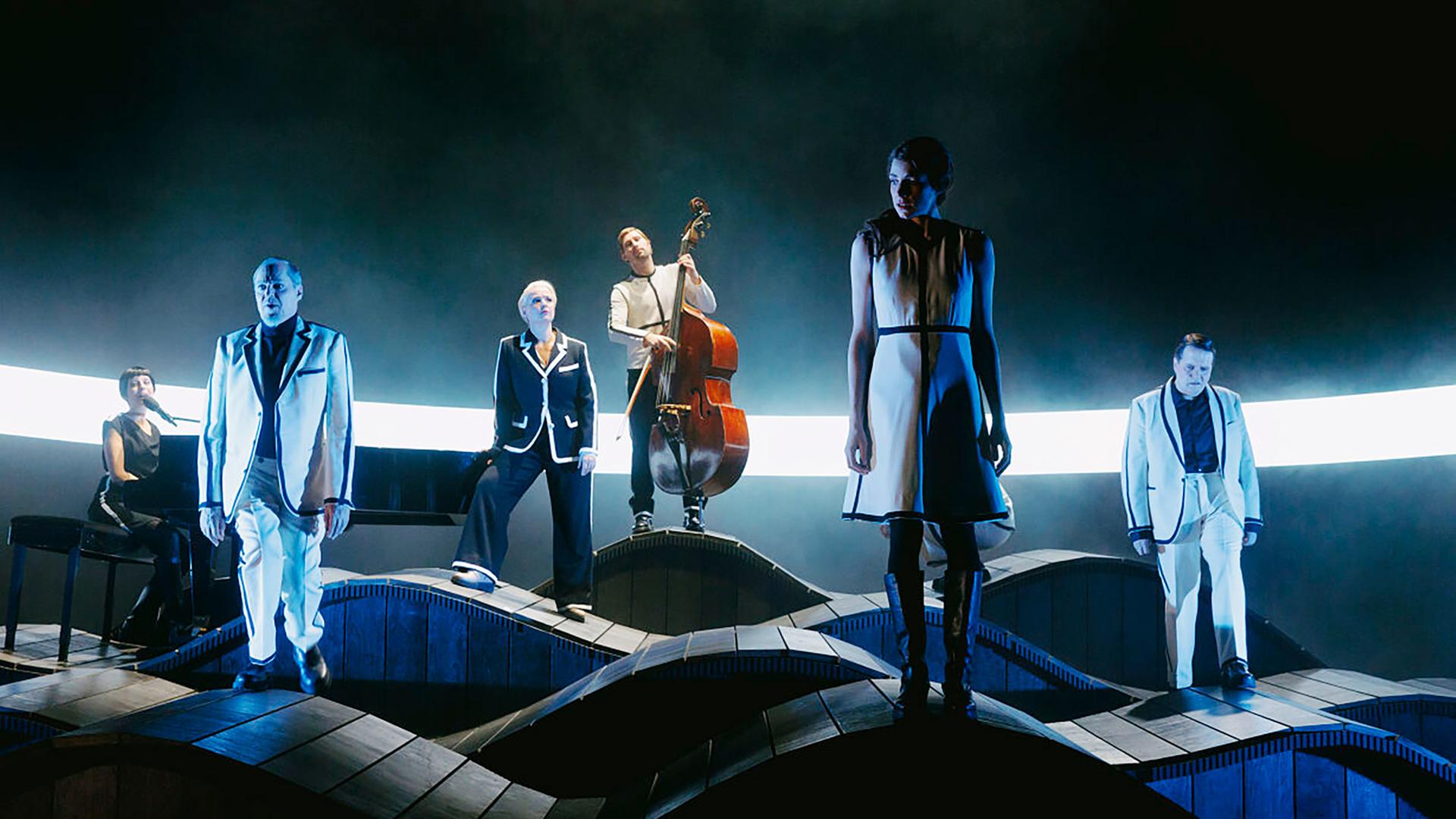 Bühnenszene: In einer schummrigen Beleuchtung stehen Menschen auf welligem Holzboden. Ein Mann spielt Kontrabass, links spielt jemand auf einem Flügel.