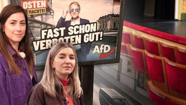 Zwei Frauen stehen vor einem Wahlplakat der Partei "AfD" (Alternative für Deutschland). Das Plakat trägt den Slogan "Fast schon verboten gut!" und zeigt einen Mann (Björn Höcke) mit Sonnenbrille, der selbstbewusst posiert. Im Hintergrund ist das Reichstagsgebäude zu sehen. Zudem steht auf dem Plakat "Der Osten macht's". Im Hintergrund des Bildes sind leere rote Theatersitze und eine Straße mit einem schwarzen Van zu sehen.
