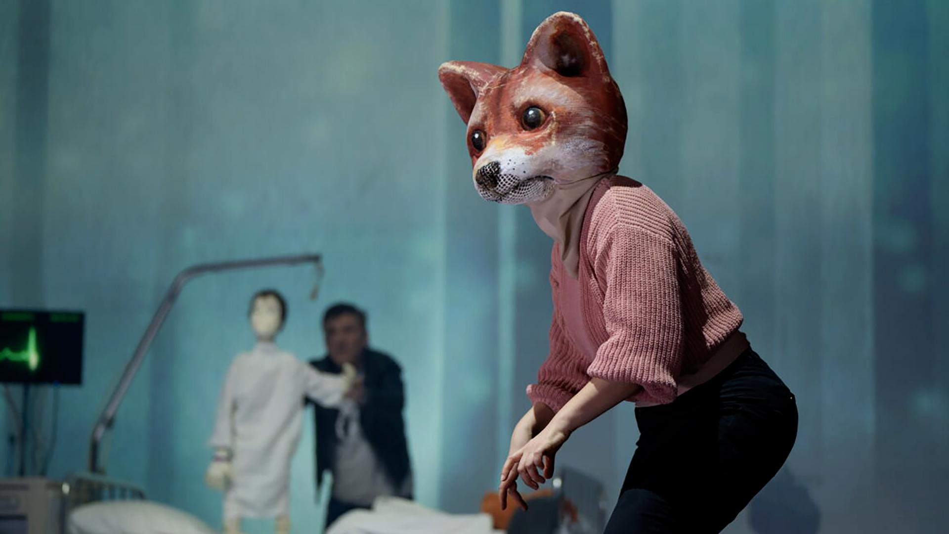 Bühnenszene: Ein Mensch, der eine große Fuchsmaske trägt, steht vor einem Krankenbett. Hinter dem Krankenbett steht ein Mann, der eine Puppe in Form eines Kindes hält.