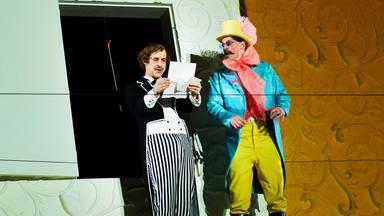 Bühnenszene: Ein Mann im Frack liest einen Brief, ein anderer Mann, der einen bunten Frack und Zylinder trägt, schaut ihn an.