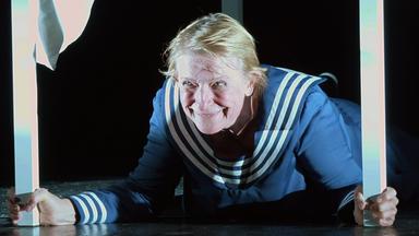 „THEATER.MACHER.INNEN“: Dagmar Manzel liegt in einem Matrosen-Kostüm und einem weißen Tisch und hält mit beiden Händen zwei Tischbeine in der Hand. Sie grinst verschmitzt nach vorne.