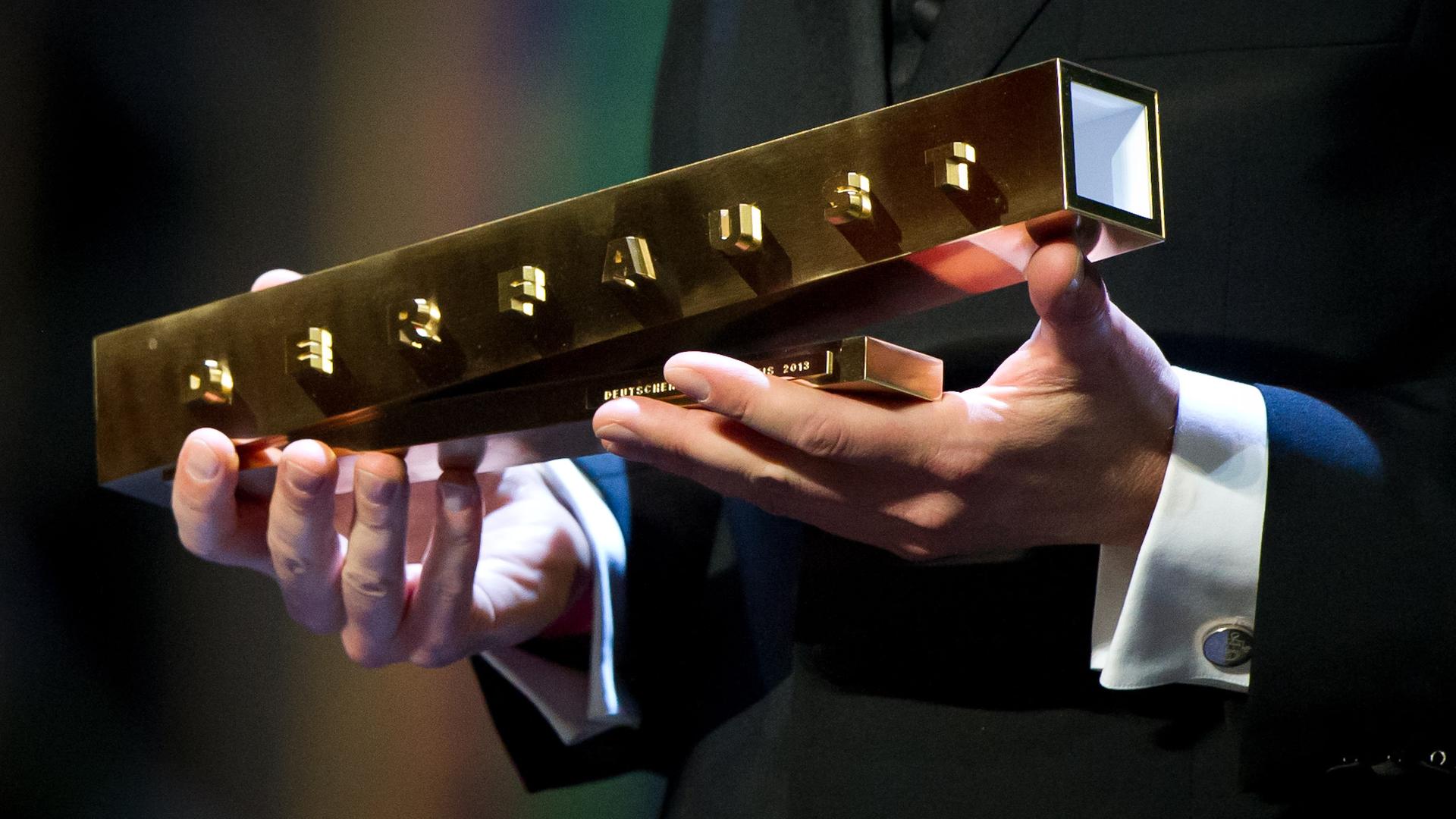 Zwei Hände halten einen goldenen Preis, auf welchem ebenfalls in goldener Farbe "der Faust" in hervorstehenden Buchstaben geschrieben steht.