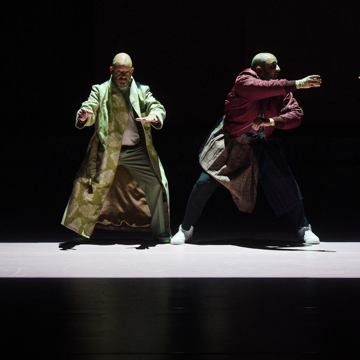 : Auf einer dunklen Bühne ist mit Licht auf dem Boden ein Rechteck abgegrenzt. Links an der schmalen Seite steht eine Frau mit auf Höhe der Hüfte erhobenen Armen, sie trägt ein glänzendes, buntgemustertes langes Kleid. Rechts neben ihr, nach vorne gewandt, ebenfalls mit erhobenen Armen steht mit leicht gespreizten Beinen ein glatzköpfiger Mann, er trägt einen langen hellgrünen Mantel. Neben ihm in ähnlicher Position ein weiterer, etwas jüngerer glatzköpfiger Mann, in einer dunkelroten Bomberjacke, einem Schottenrock über der Jeans und weißen Turnschuhen. Er wendet sich nach rechts, dort steht an der anderen Schmalseite eine junge blonde Frau in einem weißen Kleid, mit Blümchenmuster. Sie streckt den rechten Arm zu dem Mann neben ihr aus. Zu ihren Füßen liegt rechts ein Football. 