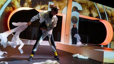 Eine mit Projektionen bespielte Wand, mit zwei an den Seiten abgerundeten Öffnungen, in der Mitte eine Art Laufsteg. Links davon im Bildvordergrund eine Frau in Bewegung, die einen weißes Tuch hinter sich wirft, um sie herum liegen weitere weiße Tücher. Im Hintergrund zwei Männer. Einer trägt schwarz und hält ein Stück Tüll in der Hand, der andere am rechten Bildrand, mit dem Rücken zum Publikum, eine weiße Bomberjacke. 