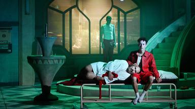 Düstere Theaterszene mit expressionistischem Bühnenbild: Eine Person in einem roten Hemd und roter Hose sitzt barfuß auf einem schlichten Metallbett, mit leerem Blick nach vorne. Eine andere Person in weißem Hemd und dunkler Hose liegt mit dem Kopf auf ihrem Schoß, die Augen geschlossen. Im Hintergrund steht eine weitere Person mit weißem Unterhemd und roter Hose vor einem großen, schlüsselförmigen Fenster, durch das eine nächtliche Stadtszene mit Autos und Straßenlaternen zu sehen ist. Links im Bild ein Brunnen, der Boden ist gefliest. 