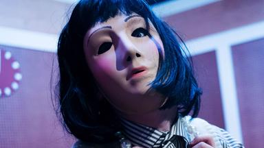 Links im Vordergrund eine Frau mit einer Silikonmaske, die ihr Gesicht vollständig verdeckt. Sie trägt eine schwarze Perücke, einen grau-weiß gestreifte Bluse und eine helle Strickweste. Rechts ein Mann, ebenfalls mit Maske und schwarzer Perücke, er trägt eine Brille und einen grauen Anzug. Zwischen ihnen ein Tisch mit brauner Linoleumtischdecke, darauf zwei umgestürzte Gläser und ein Aschenbecher.  