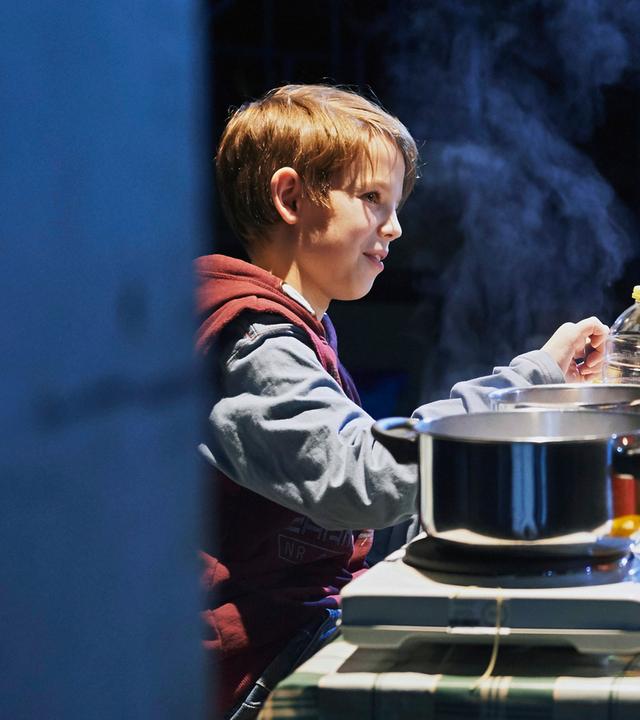 : An einem Tisch sitzt links ein kleiner Junge mit blonden, kurzen Haaren, ihm gegenüber eine Frau mit langen, hellbraunen Haaren. Sie blicken sich an. Die Frau hat den linken Ellbogen aufgestützt, sie isst Spaghetti. Auf dem Tisch steht eine Kochplatte mit zwei Feldern, darauf zwei Töpfe, aus dem größeren im Vordergrund steigt Dampf auf.  Auf dem Tisch stehen außerdem eine rote Thermoskanne, eine kleine gelbe Plastikschüssel und im Hintergrund eine Ölflasche. 