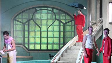 Theaterszene in einem stilisierten, expressionistischen Badehaus: Zwei Schauspieler in roten Hosen stehen vorn, einer mit weißem Unterhemd, der andere mit rotem Hemd. Sie halten sich an den Händen und lächeln. Im Hintergrund steht ein Mann in roter Kleidung mit einem schwarzen Regenschirm auf einer Treppe, die zu einem runden Wasserbecken führt. Links ist eine Person in einer schmutzigen Schürze zu sehen, die an einem Metallwagen arbeitet. Drei nackte Körper liegen regungslos auf dem gefliesten Boden. Das Bühnenbild ist geprägt von grünen Jugendstilfenstern, runden Wandverzierungen und einer unheimlichen Atmosphäre.