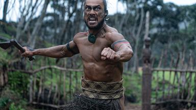 Ein Maori mit Streitaxt stürmt mit weit aufgerissenen Augen und wütendem Gesichtsausdruck auf die Betrachter zu.