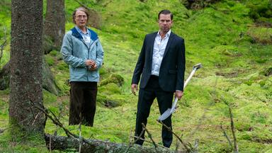 Zwei Männer stehen im Wald. Einer trägt einen Anzug und hält eine Schaufel in der linken Hand. Der zweite steht etwas verzagt an seiner Seite.