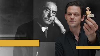 Jo Schück hält eine Playmobil Mann Figur, mit Thomas Mann im Hintergrund