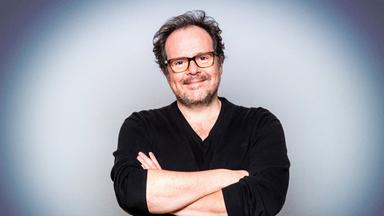 Das Bild zeigt einen Mann mittleren Alters mit einem freundlichen Gesichtsausdruck, der leicht lächelt. Er hat dunkle, lockige Haare mit einigen grauen Strähnen und trägt eine Brille mit einem rechteckigen Rahmen. Sein Outfit besteht aus einem schwarzen, offenen Pullover, der über einem schwarzen T-Shirt getragen wird.   Der Mann steht mit verschränkten Armen vor einem sanft verlaufenden, helleren Hintergrund, der von einem blassblauen bis weißen Farbton reicht. Die Beleuchtung ist gleichmäßig und sorgt für eine angenehme Darstellung des Gesichts und der Kleidung. Das Bild vermittelt eine freundliche und entspannte Atmosphäre.