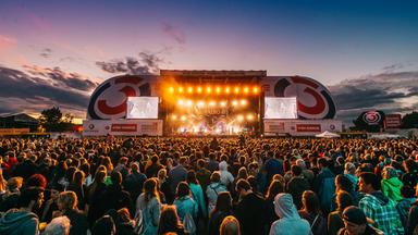 Das Bild zeigt eine bunte und feierliche Atmosphäre am Donauinselfest in Wien. Auf der Bühne befinden sich große, hell erleuchtete Scheinwerfer, die ein warmes Licht ausstrahlen. Zu sehen sind auch zwei große Leinwände, auf denen möglicherweise Auftritte oder Videos gezeigt werden. Im Vordergrund steht eine große Menschenmenge, die dicht gedrängt vor der Bühne steht und die Performance anscheinend enthusiastisch verfolgt. Die Zuschauer sind in einer Vielzahl von Outfits gekleidet, die von lässig bis festlich reichen. Der Himmel ist zur Dämmerungszeit gefärbt und zeigt sanfte Farbverläufe von Blau bis Orange, was zur festlichen Stimmung beiträgt. Aus verschiedenen Richtungen sind Zelte und Stände zu erkennen, die wahrscheinlich zusätzliche Aktivitäten oder Verkaufsstände darstellen.
