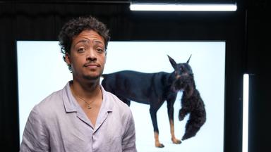 Ein Mann in hellem Hemd steht in einem dunklen Raum, hinter ihm sieht man ein Foto eines Dobermanns, der einen Pudel im Maul trägt. Der Mann legt die Stirn in Falten. 