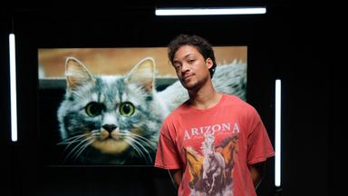 Ein Mann steht vor einem großen Foto einer Katze mit grünen Augen.  Er trägt ein rotes Shirt mit Pferden darauf.  