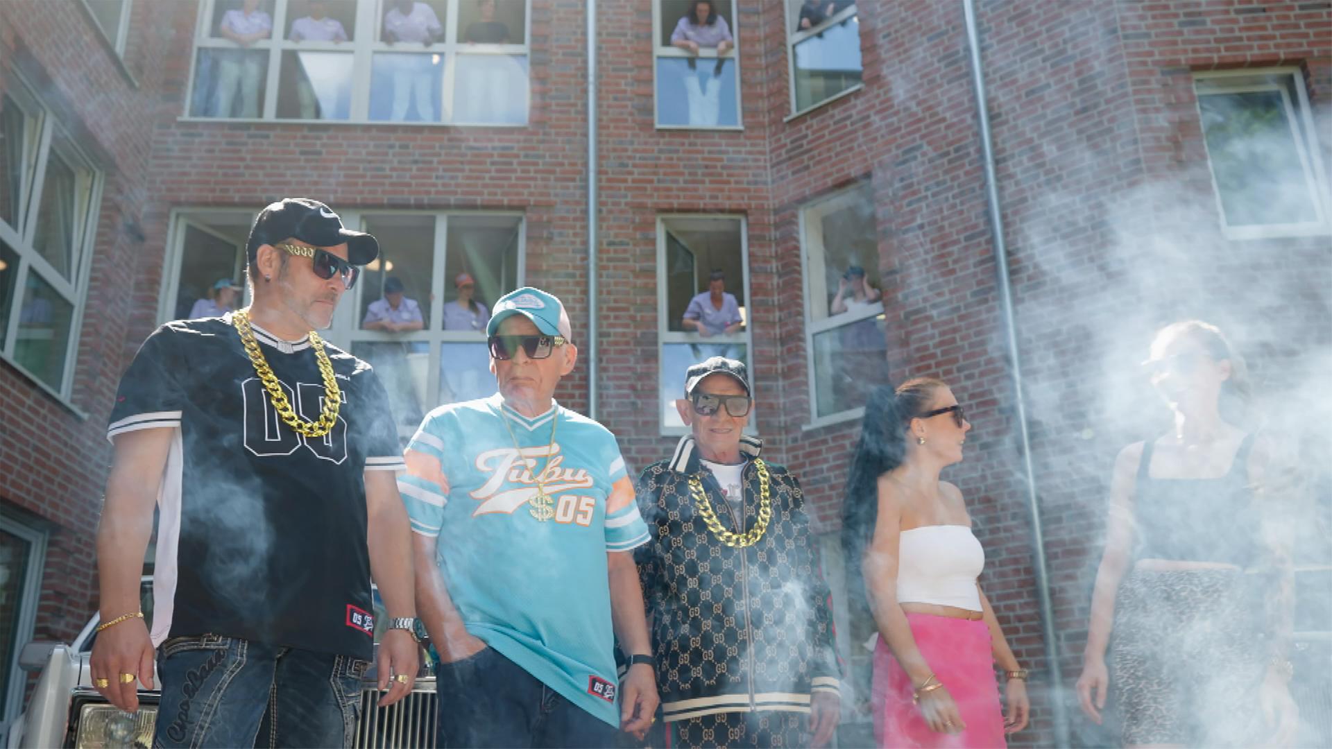 Drei wie Gangster-Rapper gekleidete Männer und zwei junge Frauen in engen Tops stehen vor einem Haus, in dessen Fenster jeweils eine Person steht und nach draußen schaut.