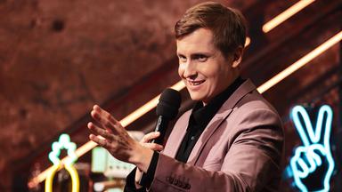 Till Reiners steht auf der rechten Seite des Bildes.Er lacht und hält in seiner rechten Hand ein schwarzes Mikrofon. Seine linke Hand streckt er nach vorne