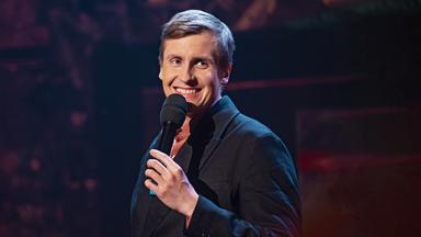 Till Reiners steht auf der rechten Seite des Bildes. Er lacht und hält in seiner linken Hand ein schwarzes Mikrofon. 