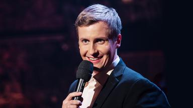 Till Reiners steht auf der rechten Seite des Bildes. Er lacht und hält in seiner linken Hand ein schwarzes Mikrofon.