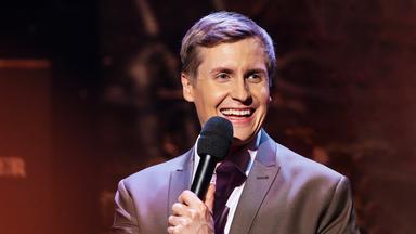 Till Reiners steht auf der rechten Seite des Bildes. Er lacht und hält in seiner linken Hand ein schwarzes Mikrofon