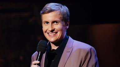 Till Reiners steht auf der rechten Seite des Bildes. Er lächelt und hält in seiner Hand ein schwarzes Mikrofon.