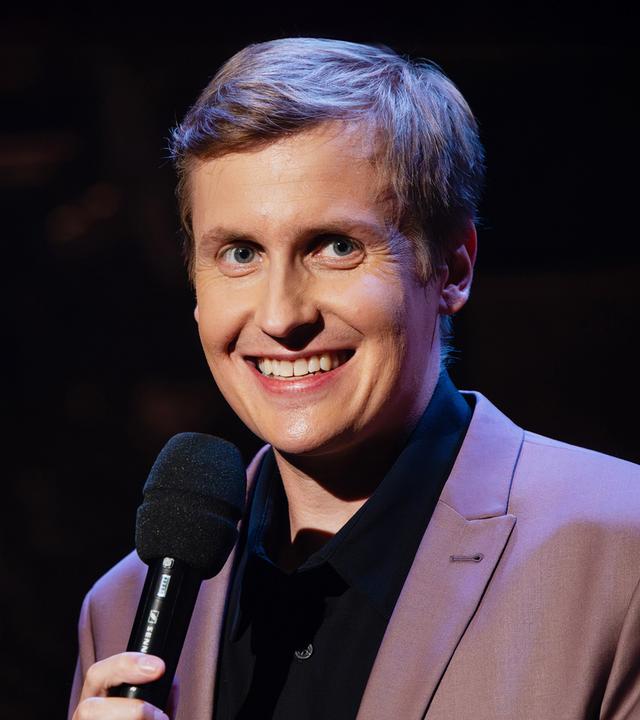 Till Reiners steht auf der rechten Seite des Bildes. Er lächelt und hält in seiner Hand ein schwarzes Mikrofon.