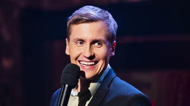 Till Reiners ist in der Mitte des Bildes zu sehen. Vor seinem Kinn ist ein Mikrofon. Der Hintergrund ist dunkel. Till Reiners lächelt.