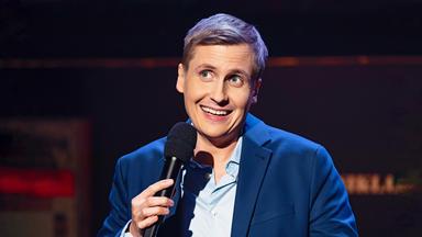 Till Reiners ist in der Mitte des Bildes zu sehen. Till Reiners lächelt und schaut in Richtung der linken Bildecke. In seiner rechten Hand hält er ein Mikrofon.