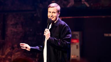 Till Reiners ist auf der linken Bildseite zu sehen. Till Reiners lächelt und schaut in Richtung der rechten Bildseite. In seiner linken Hand hält er ein Mikrofon. 