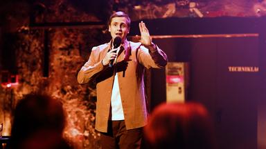 Till Reiners in der Mitte des Bildes. In seiner rechten Hand hält er ein Mikrofon. Die Beleuchtung ist rot-orange.