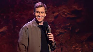 Till Reiners ist auf der rechten Bildseite zu sehen. Till Reiners lächelt und schaut in Richtung der linken Bildseite. In seiner linken Hand hält er ein Mikrofon. Die Beleuchtung ist rot und lila.