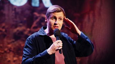 Till Reiners ist auf der rechten Bildseite zu sehen. Er greift sich mit seiner linken Hand an die Stirn. In der rechten Hand hält er sein Mikrofon.