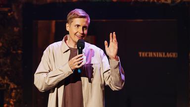 Till Reiners hält ein Mikrofon in einer Hand, mit der anderen Hand gestikuliert er. Er trägt ein braunes Shirt und darüber eine beige Jacke.