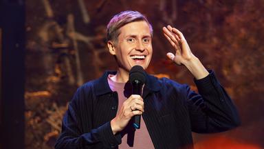 Till Reiners ist auf der rechten Bildseite zu sehen. In seiner rechten Hand hält er ein Mikrofon und hebt dazu den linken Arm auf Kopfhöhe an. Er lächelt.