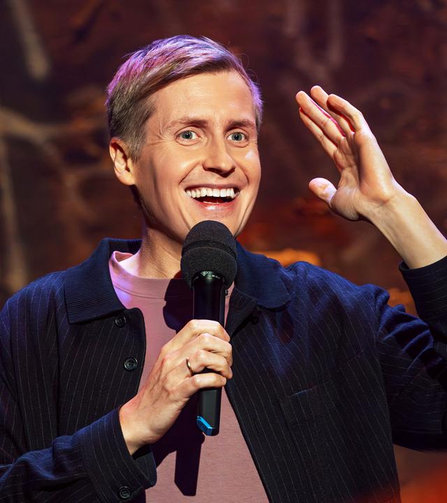 Till Reiners ist auf der rechten Bildseite zu sehen. In seiner rechten Hand hält er ein Mikrofon und hebt dazu den linken Arm auf Kopfhöhe an. Er lächelt.