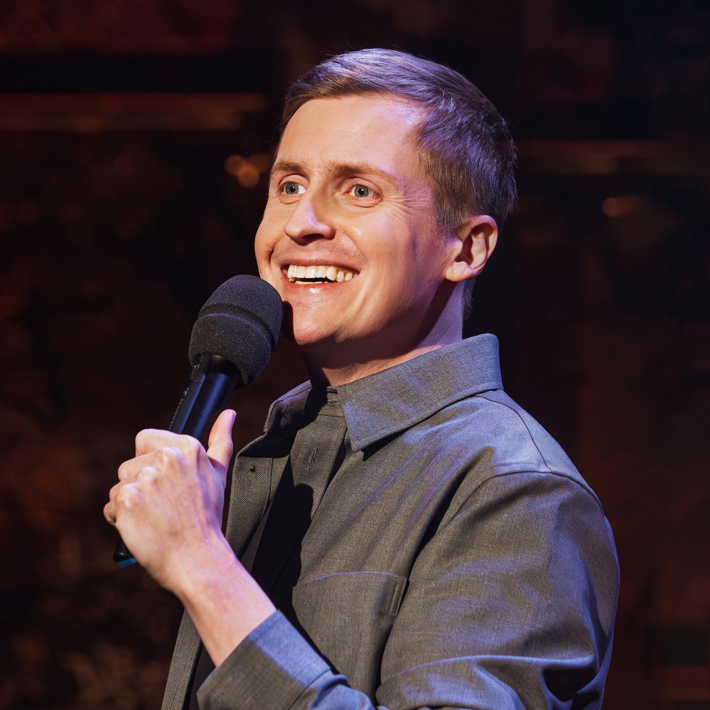 Till Reiners ist auf der rechten Bildseite zu sehen. In seiner linken Hand hält er ein Mikrofon. Der Hintergrund ist dunkel, sein Hemd ist grau.