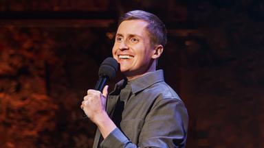 Till Reiners ist auf der rechten Bildseite zu sehen. In seiner linken Hand hält er ein Mikrofon. Der Hintergrund ist dunkel, sein Hemd ist grau.