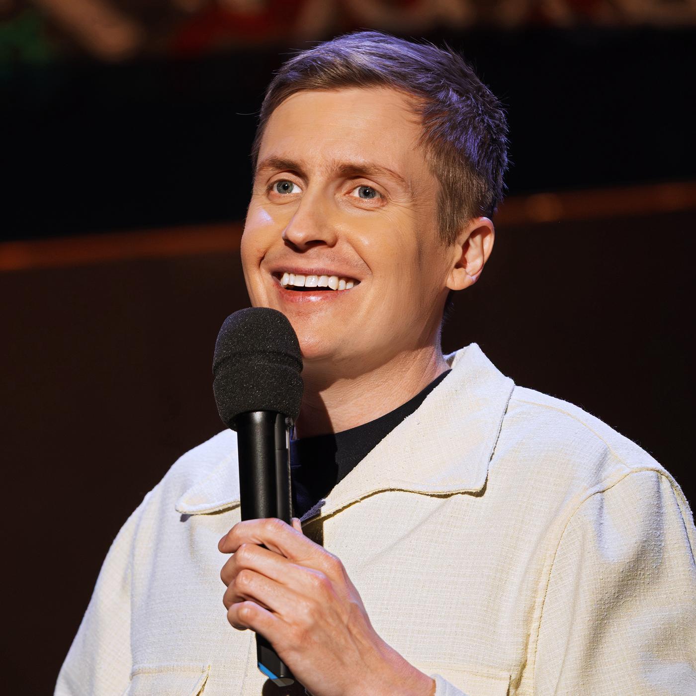 Till Reiners ist auf der rechten Bildseite zu sehen. In seiner linken Hand hält er ein Mikrofon, die rechte Hand ist nur am unteren Bildrand zu sehen. Er trägt eine offene Jacke in der Farbe beige. Er lächelt.