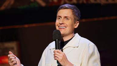 Till Reiners ist auf der rechten Bildseite zu sehen. In seiner linken Hand hält er ein Mikrofon, die rechte Hand ist nur am unteren Bildrand zu sehen. Er trägt eine offene Jacke in der Farbe beige. Er lächelt.