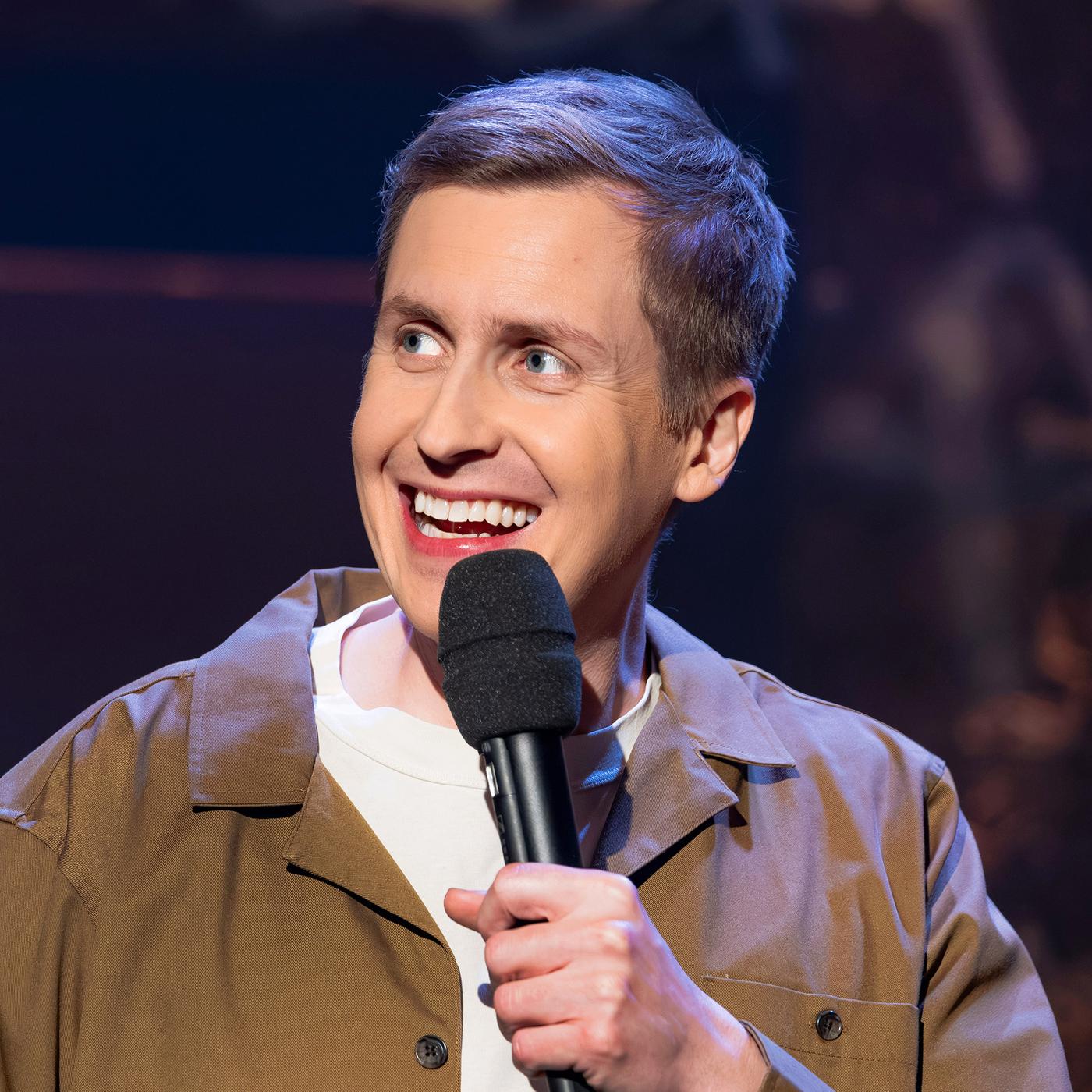 Till Reiners ist auf der rechten Bildseite zu sehen. In seiner linken Hand hält er ein Mikrofon. Er trägt eine offene Jacke in der Farbe beige. Er lächelt.