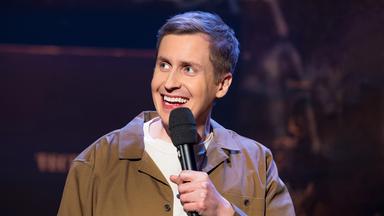 Till Reiners ist auf der rechten Bildseite zu sehen. In seiner linken Hand hält er ein Mikrofon. Er trägt eine offene Jacke in der Farbe beige. Er lächelt.