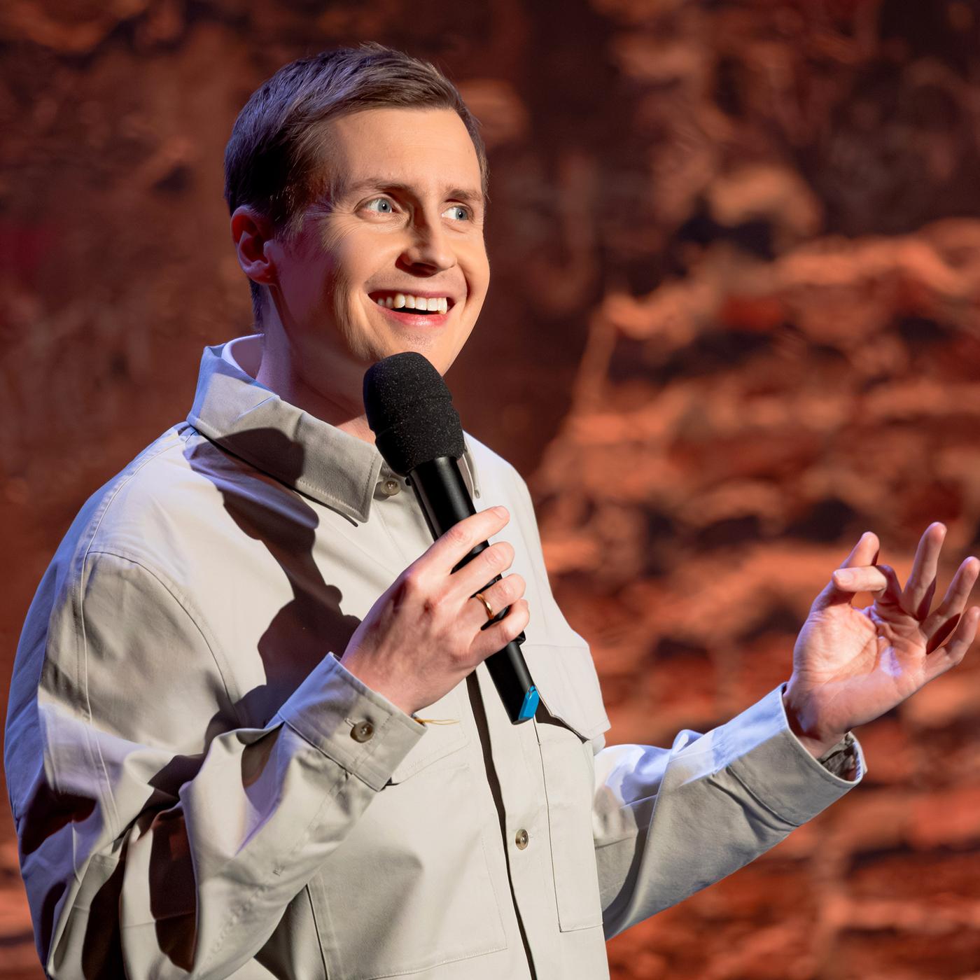 Till Reiners steht in der Mitte des Bildes. In seiner rechten Hand hält er ein Mikrofon. Er trägt ein Hemd. Till Reiners lächelt.