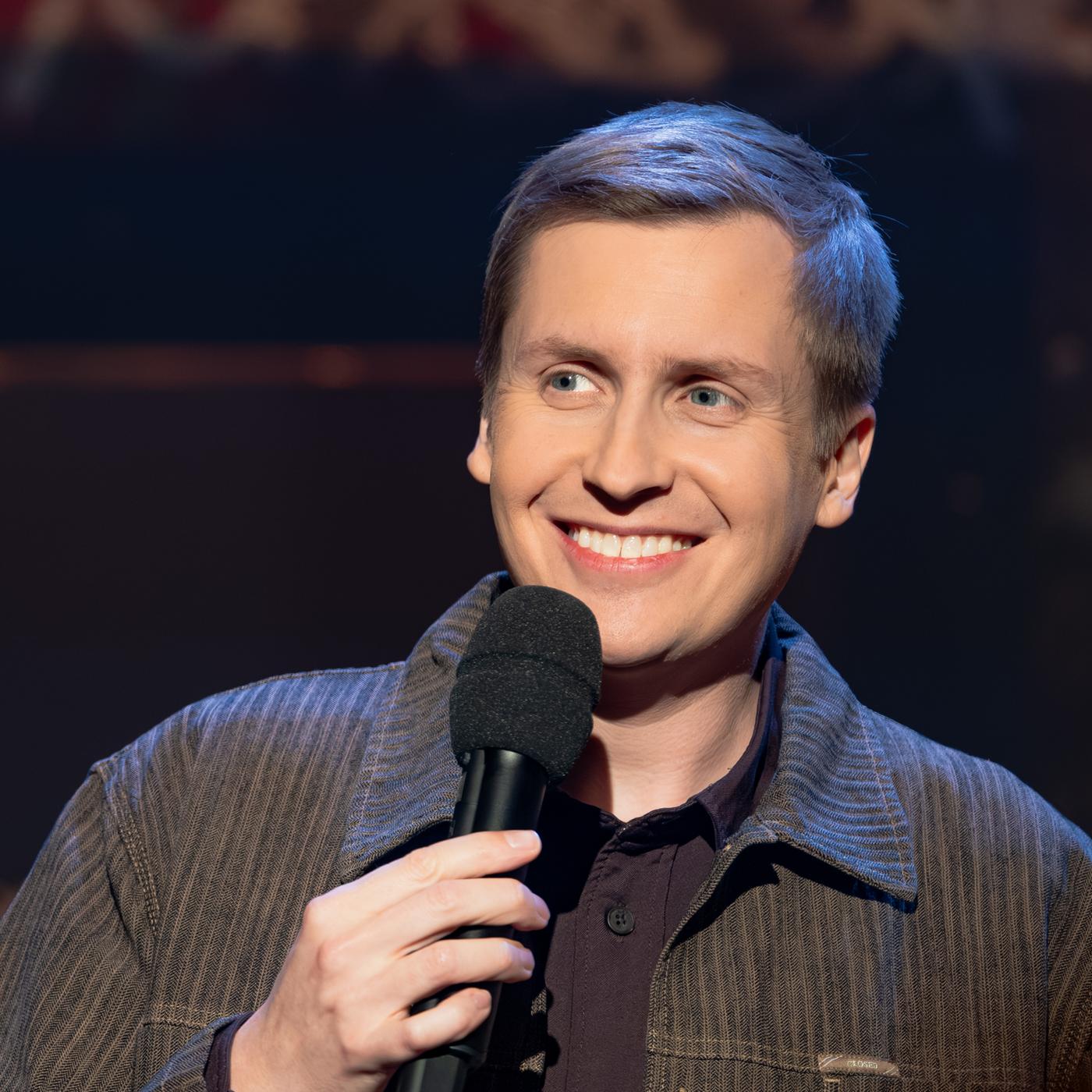 Till Reiners ist auf der rechten Seite des Bildes abgebildet. In seiner rechten Hand hält er ein Mikrofon. Reiners lächelt. Der Hintergrund ist dunkel.