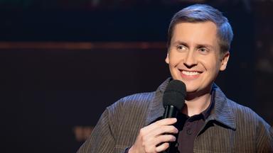 Till Reiners ist auf der rechten Seite des Bildes abgebildet. In seiner rechten Hand hält er ein Mikrofon. Reiners lächelt. Der Hintergrund ist dunkel.