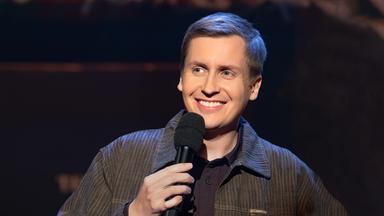 Till Reiners ist auf der rechten Seite des Bildes abgebildet. In seiner rechten Hand hält er ein Mikrofon. Reiners lächelt. Der Hintergrund ist dunkel.